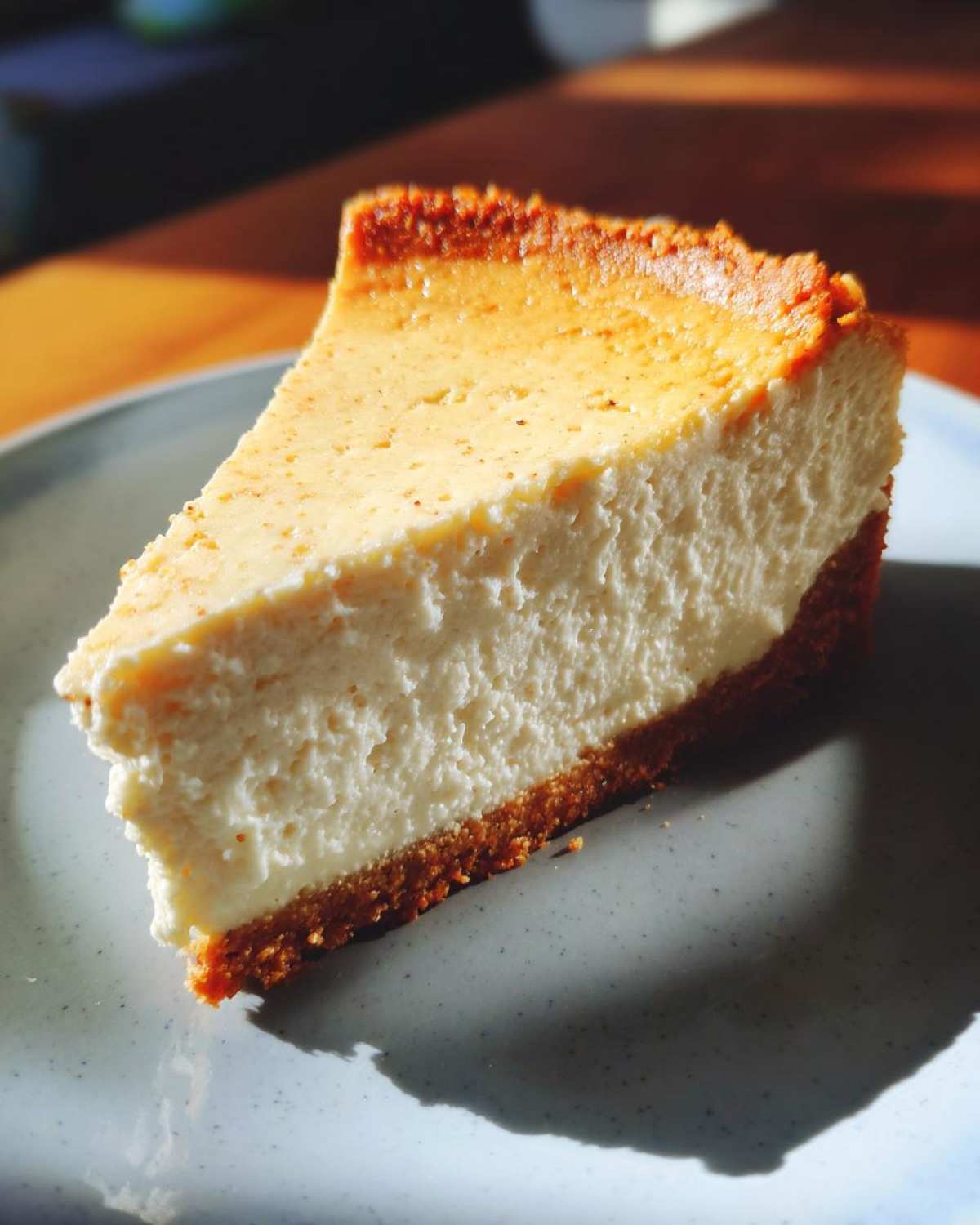 A close-up, sunlit slice of baked Protein Cheesecake showing a thick, creamy white filling and a dark graham cracker crust.