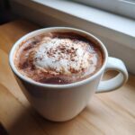 Close-up of a white mug filled with rich hot cocoa mix, topped with whipped cream and cocoa powder.