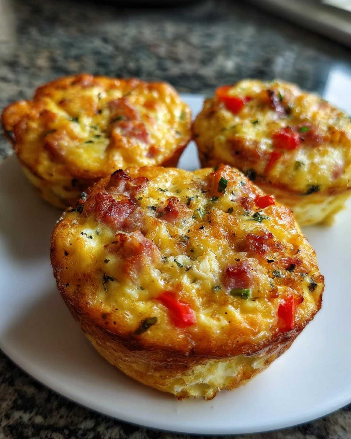 Close-up of three golden-brown savory Egg Muffin Cups topped with ham and red peppers on a white plate.