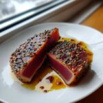 Two halves of perfectly seared tuna steak, crusted with sesame seeds, showcasing a rare, pink center, resting in marinade juices.