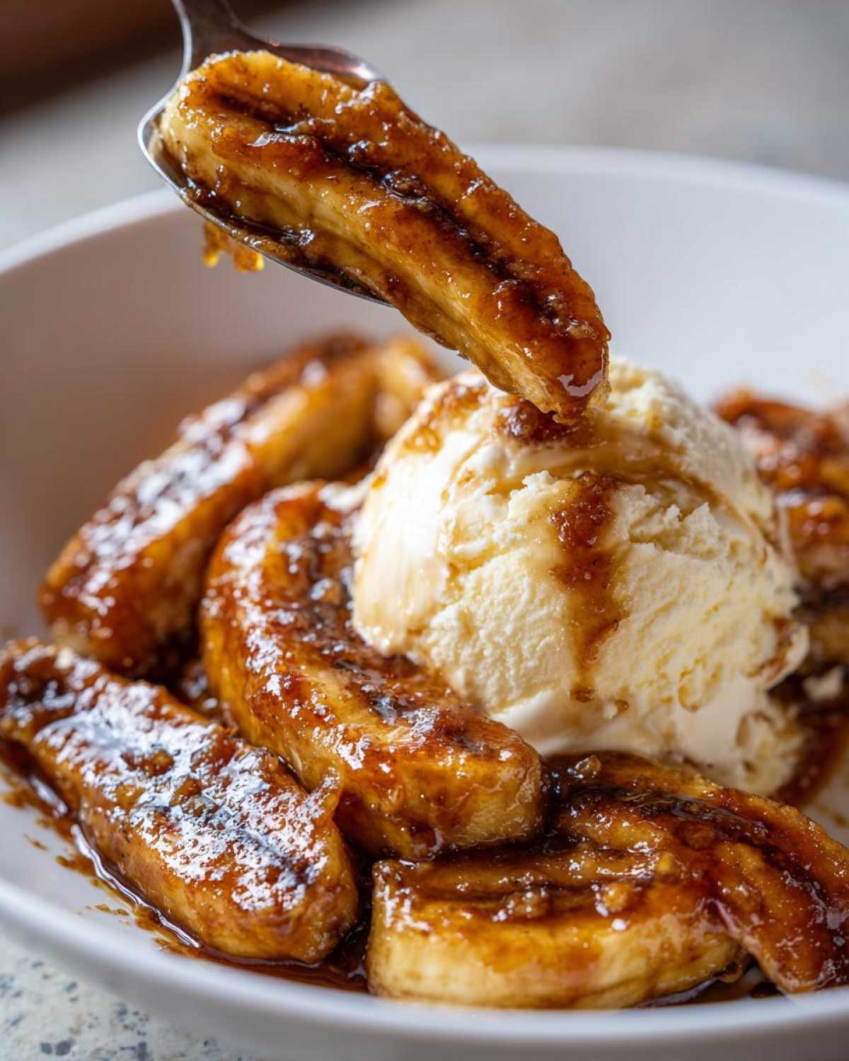 A spoonful of caramelized bananas foster being lifted from a bowl containing more bananas foster and vanilla ice cream.