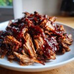 A close-up of shredded, tender Slow Cooker Pulled Pork generously coated in rich, dark BBQ sauce on a white plate.