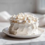 A small, round cake generously covered in fluffy vanilla buttercream frosting, sitting on a white plate.