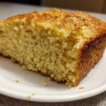 Close-up of a thick slice of golden sourdough cornbread with a shiny, sweet glaze on top, sitting on a white plate.