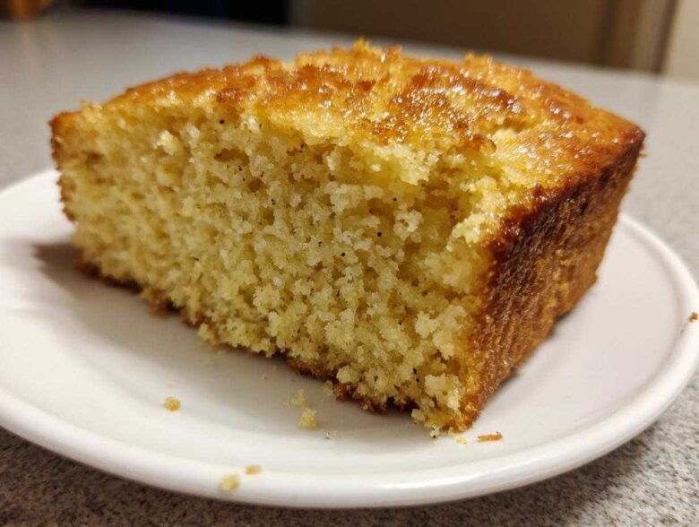 Close-up of a thick slice of golden sourdough cornbread with a shiny, sweet glaze on top, sitting on a white plate.