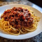 A close-up of spaghetti noodles generously topped with thick, rich bolognese sauce in a white bowl.