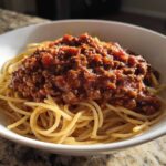 A white bowl filled with spaghetti noodles generously topped with a hearty portion of rich bolognese sauce.