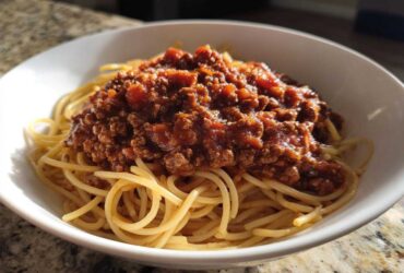 A white bowl filled with spaghetti noodles generously topped with a hearty portion of rich bolognese sauce.