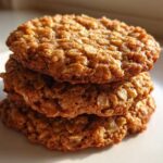 A close-up stack of three golden brown, chewy oatmeal cookie treats glistening in natural light.