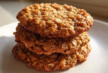 A close-up stack of three golden brown, chewy oatmeal cookie treats glistening in natural light.