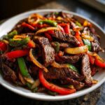 Close-up of sizzling steak fajitas with colorful strips of steak, red and green peppers, and onions piled high on a white plate.
