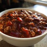 Close-up of a steaming white bowl filled with rich, hearty crockpot chili featuring ground meat and kidney beans.