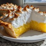 A cross-section slice of bright yellow lemon meringue pie showing the thick, glossy filling and tall, torched meringue peaks.