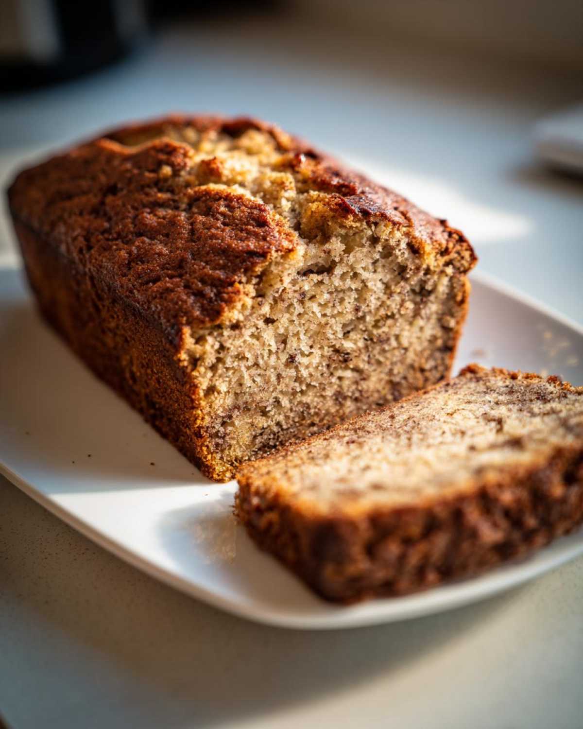 A loaf of freshly baked sugar free banana bread with one thick slice cut and resting beside it on a white platter.
