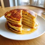 A tall stack of fluffy pancakes cut to show the airy interior, drizzled with syrup on a white plate.