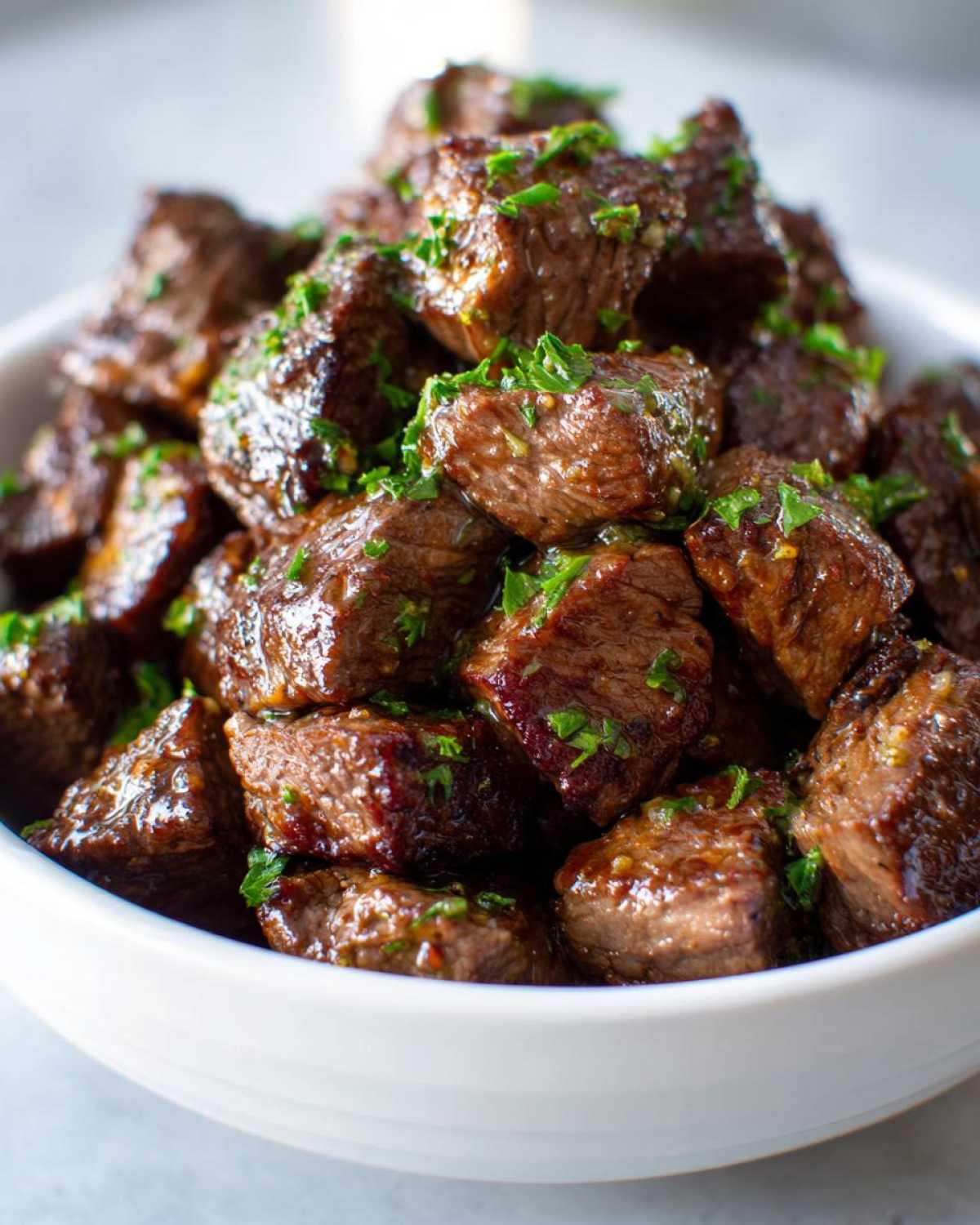 Close-up of tender, glazed crockpot steak bites piled high in a white bowl and topped with fresh chopped parsley.
