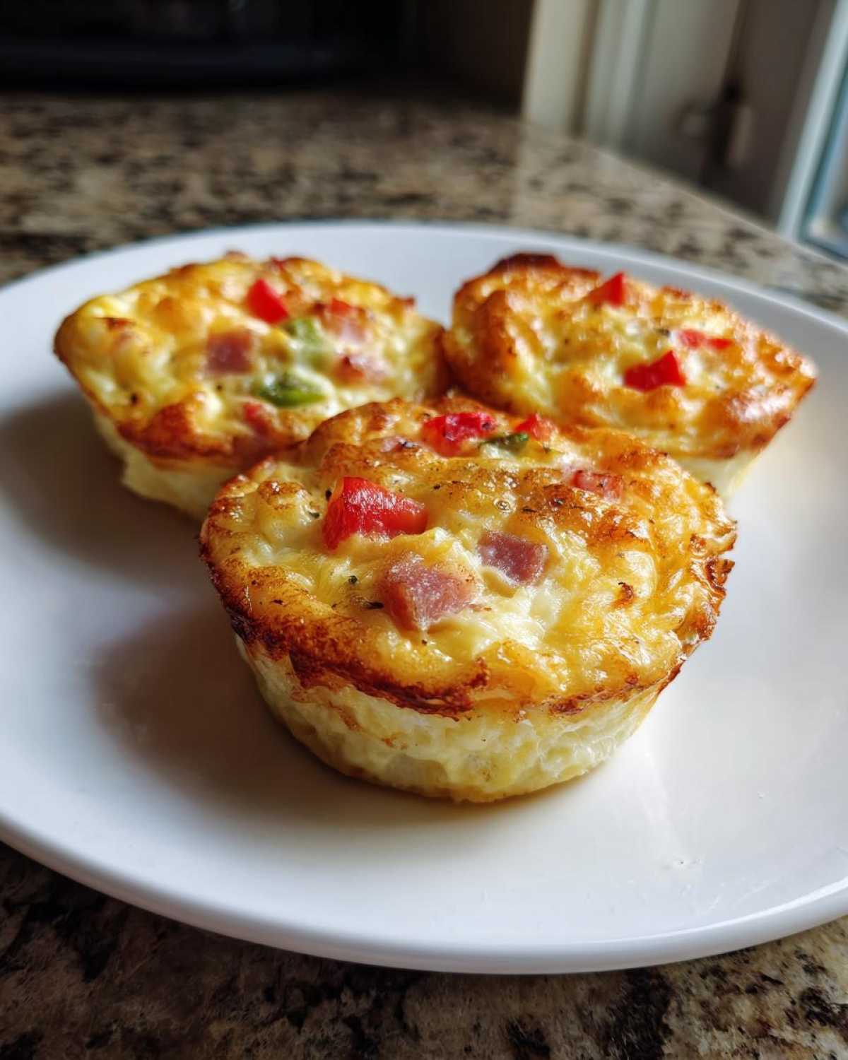 Three golden-brown Egg Muffin Cups filled with ham and colorful peppers served on a white plate.