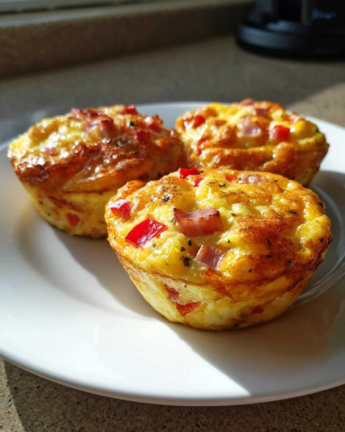 Three freshly baked Egg Muffin Cups with visible chunks of ham and red pepper, resting on a white plate.