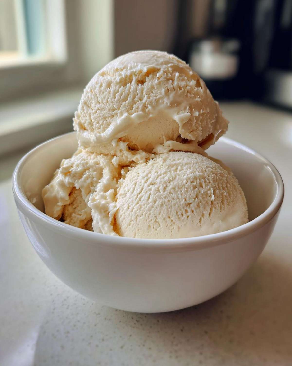 Three scoops of creamy vanilla protein ice cream served in a white bowl on a speckled countertop.