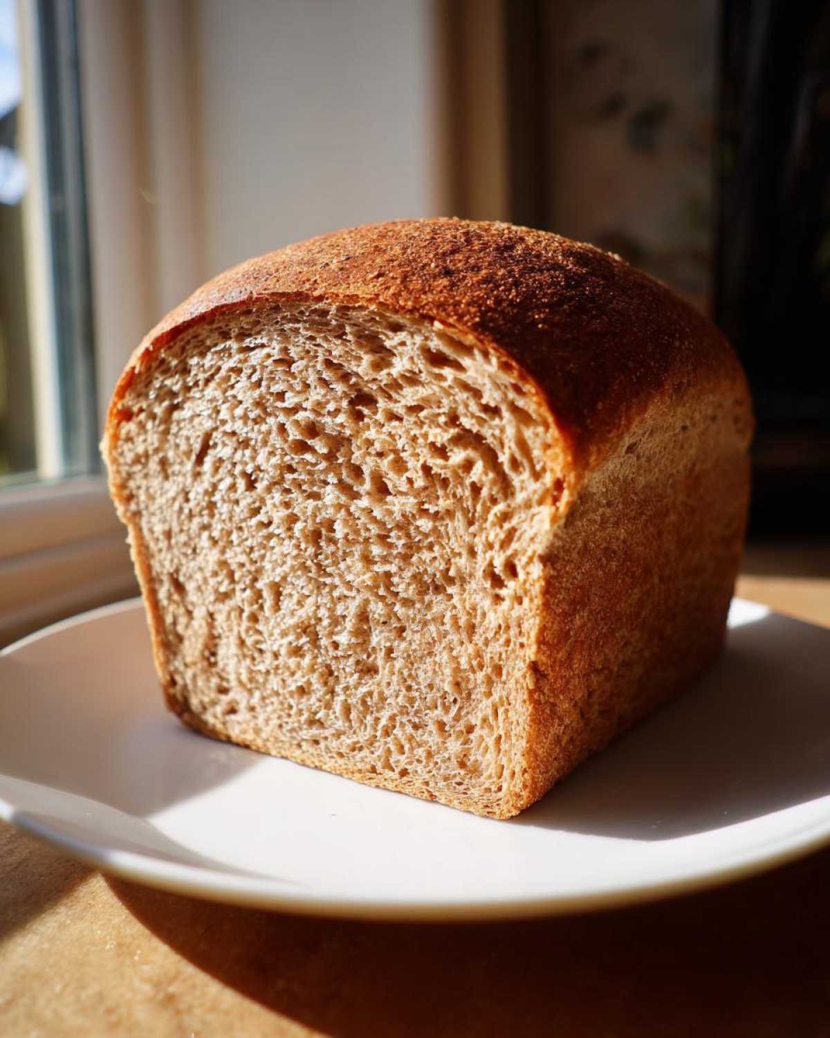 A freshly baked, square loaf of whole wheat bread, cut to show the soft crumb, resting on a white plate near a window.