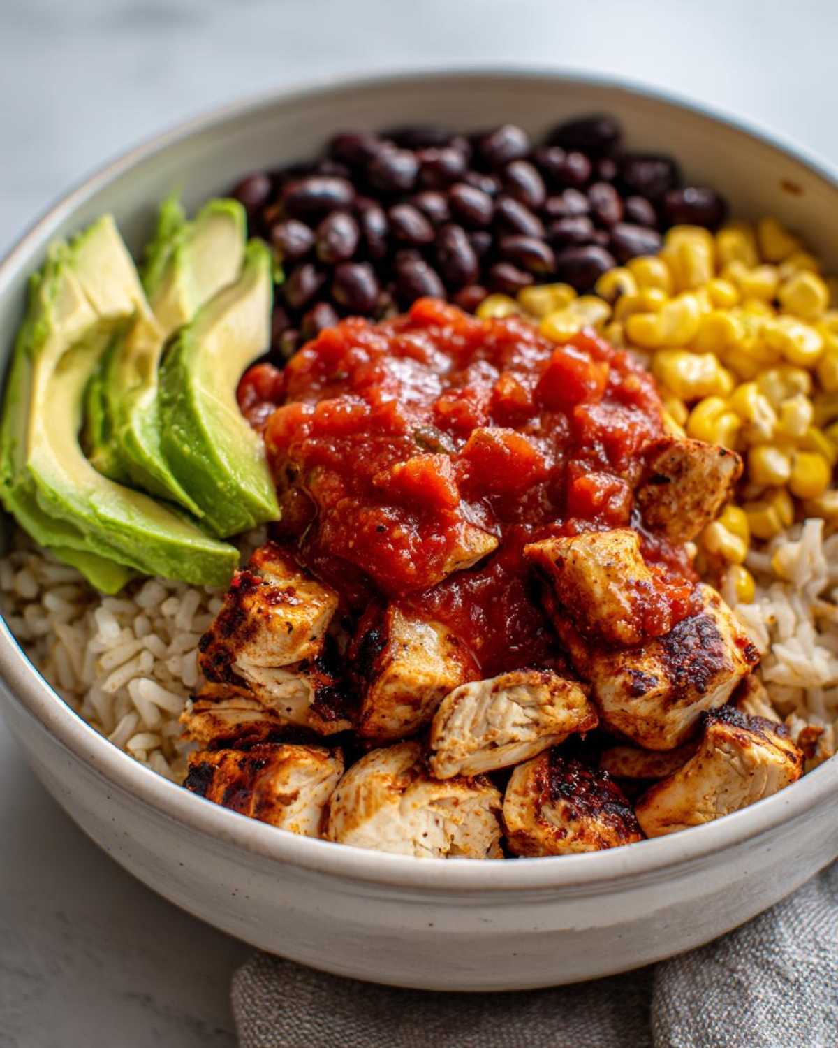 A close-up of a delicious chicken burrito bowl featuring seasoned chicken, rice, salsa, corn, black beans, and avocado slices.