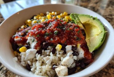 Close-up of a vibrant chicken burrito bowl featuring rice, black beans, corn, salsa, sour cream, and avocado slices.