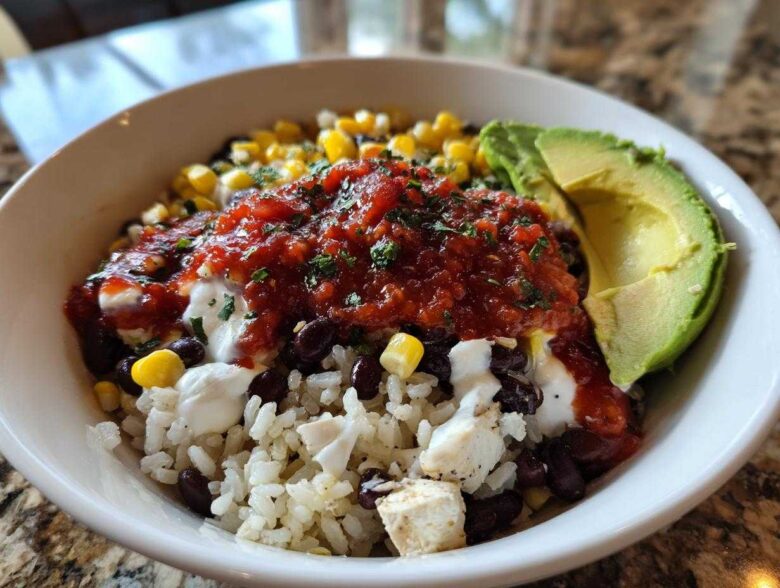 Close-up of a vibrant chicken burrito bowl featuring rice, black beans, corn, salsa, sour cream, and avocado slices.