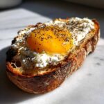Close-up of a delicious egg toast featuring a runny yolk, white cheese spread, and everything bagel seasoning.