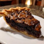 A rich, dark slice of fudge pie with a flaky crust, topped with melted chocolate chips, served on a white plate.