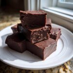 A stack of rich, dark chocolate fudge squares showing a smooth, dense texture on a white plate.
