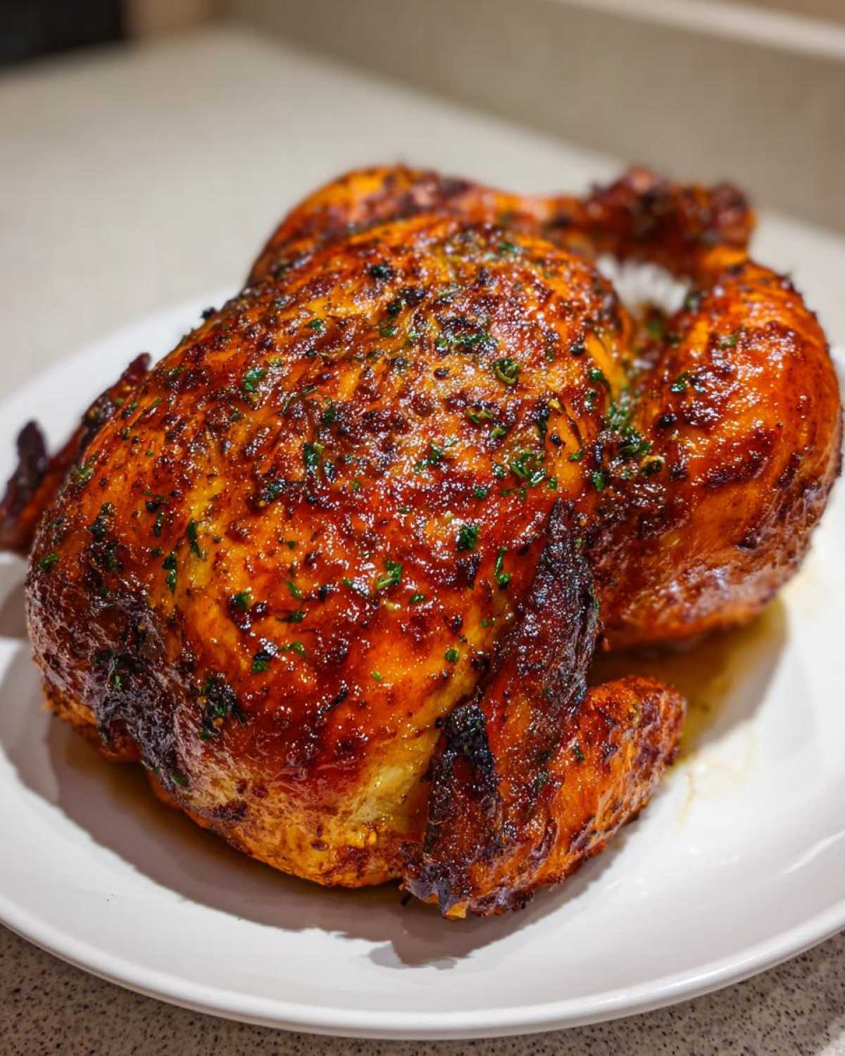 A whole, perfectly roasted chicken with deeply browned, glazed skin sprinkled with herbs, resting on a white plate.
