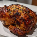 A perfectly cooked, whole roasted chicken with glossy, dark brown, crispy skin seasoned with herbs.