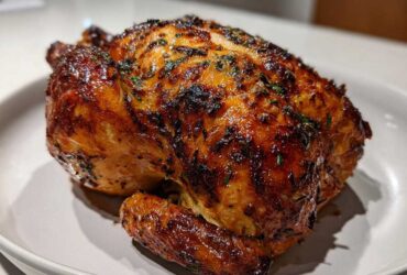 A perfectly cooked, whole roasted chicken with glossy, dark brown, crispy skin seasoned with herbs.