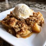 A warm slice of apple crisp recipe topped with a melting scoop of vanilla ice cream on a white plate.
