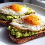 Close-up of two slices of avocado toast topped with mashed avocado and perfectly fried eggs sprinkled with seasoning.