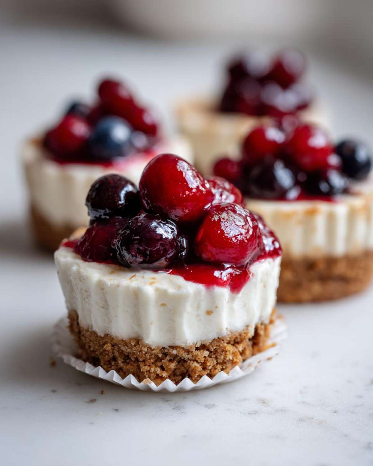 A close-up of a single mini cheesecake topped with glossy red and dark berries in sauce.