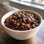 A close-up of a white bowl filled with thick, rich black bean chili, set on a dark wooden surface.
