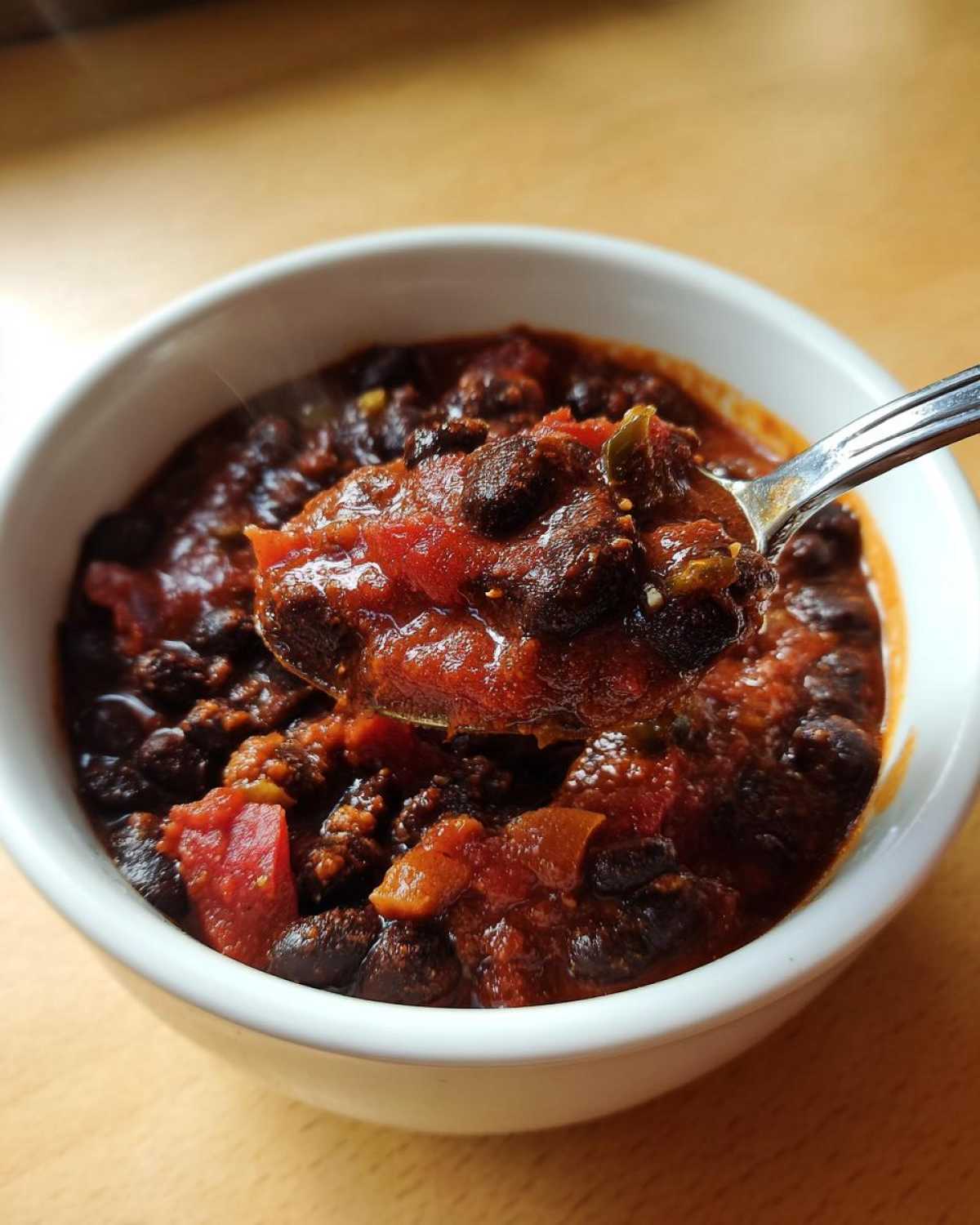 A spoonful of rich, steaming black bean chili lifted from a white bowl, showing chunks of tomato and black beans.