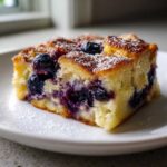 Close-up of a square slice of blueberry french toast casserole, dusted with powdered sugar, on a white plate.