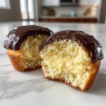Close-up of a boston cream pie cupcake cut in half, showing vanilla cake, pastry cream filling, and chocolate ganache topping.