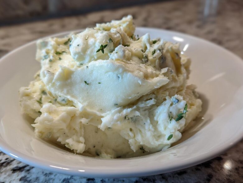 Close-up of creamy Boursin mashed potatoes mixed with herbs served in a white bowl.