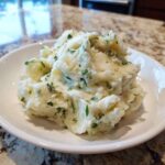 A serving of creamy Boursin mashed potatoes mixed with visible green herbs in a white bowl.