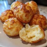 A pile of golden-brown brazilian cheese bread puffs on a plate, one broken open showing the soft, cheesy interior.