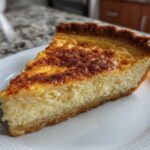 A close-up of a thick slice of buttermilk pie on a white plate, showing its creamy texture and caramelized top.