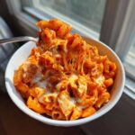 A fork lifting a cheesy, saucy bite of enchilada pasta out of a white bowl.