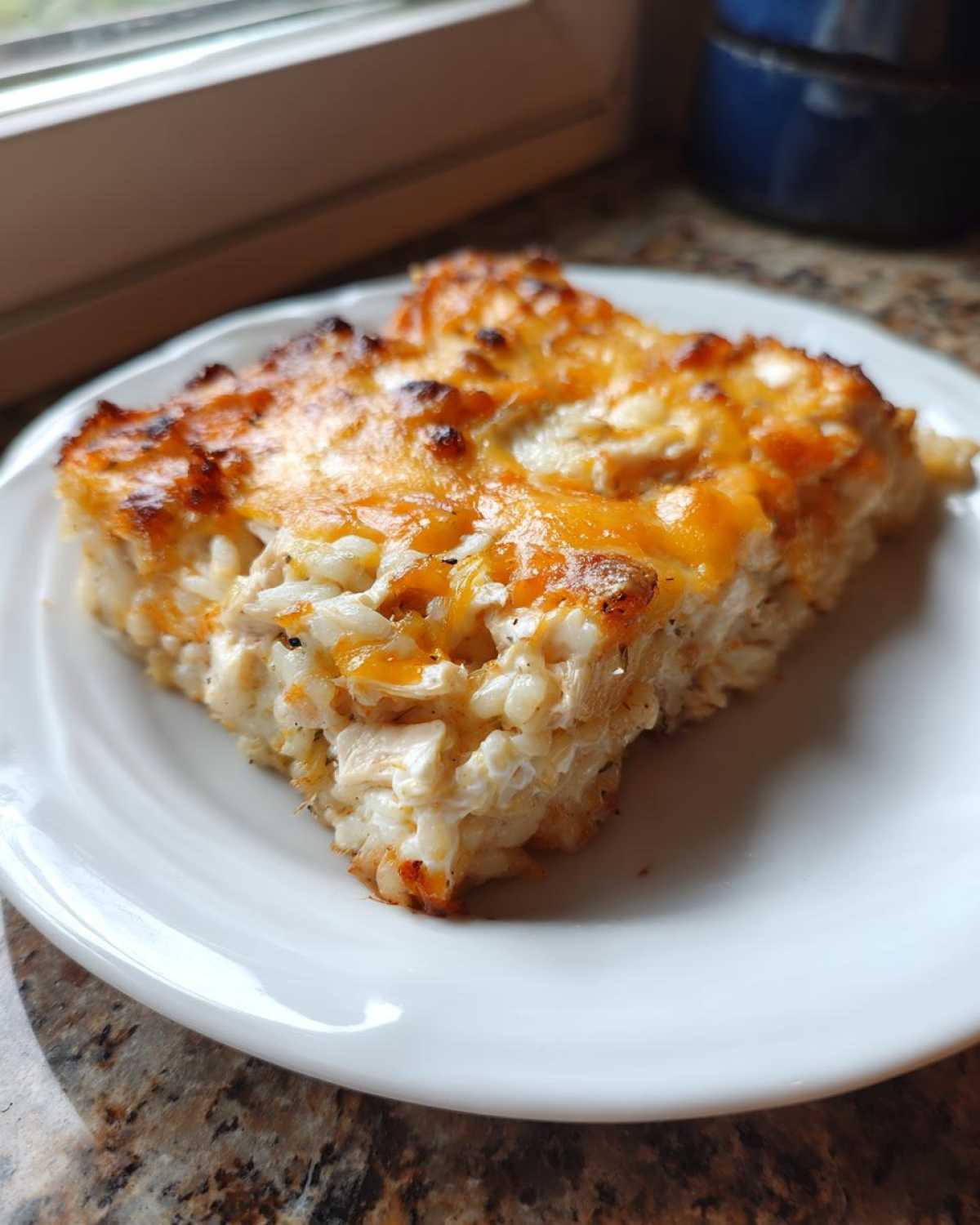 A square slice of creamy, cheesy rice casserole with a golden-brown, baked top, served on a white plate.