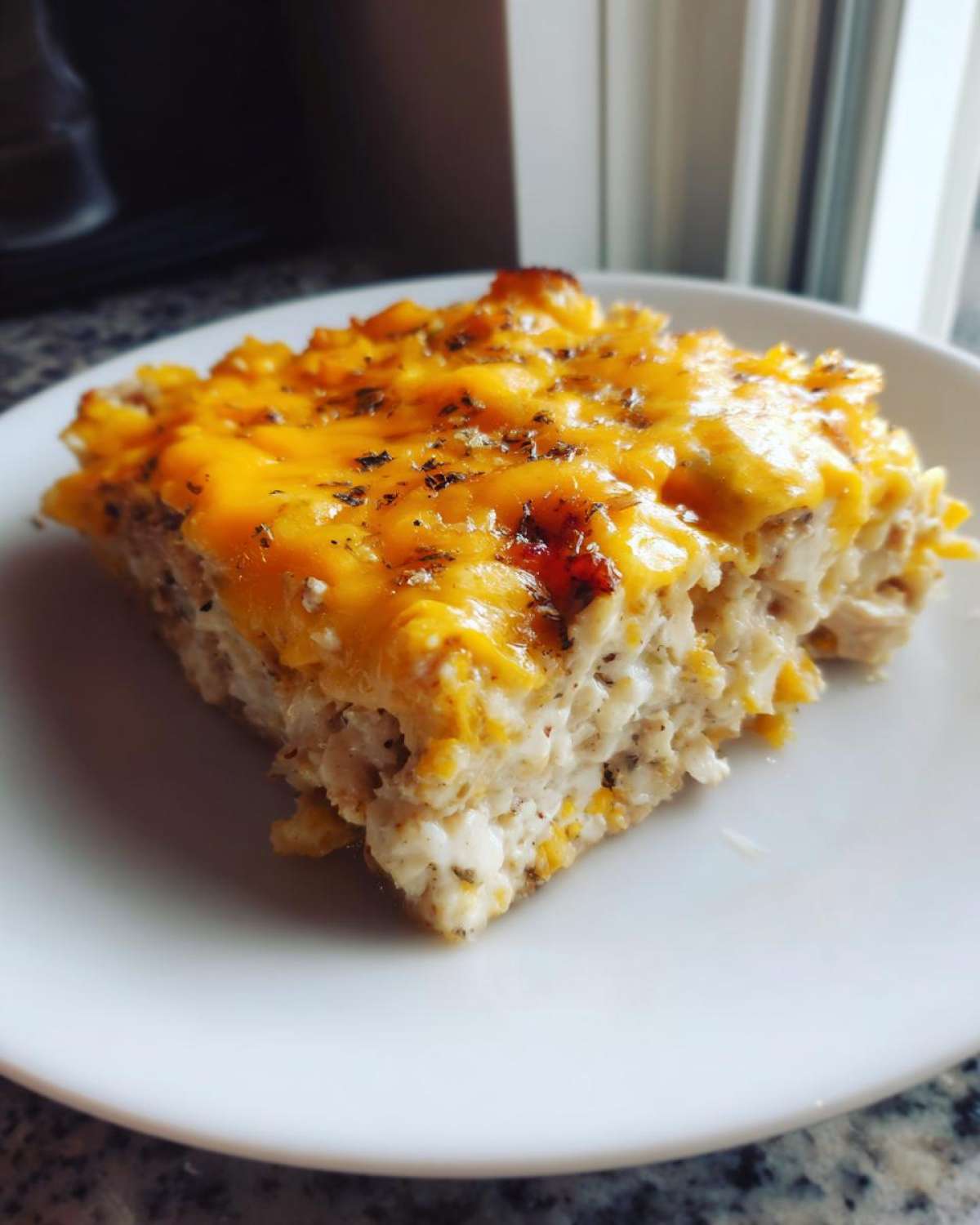 A square serving of creamy rice casserole topped with melted cheddar cheese and herbs.