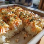A close-up view of a freshly baked sushi bake with a golden, cheesy top and visible layers of rice and salmon.