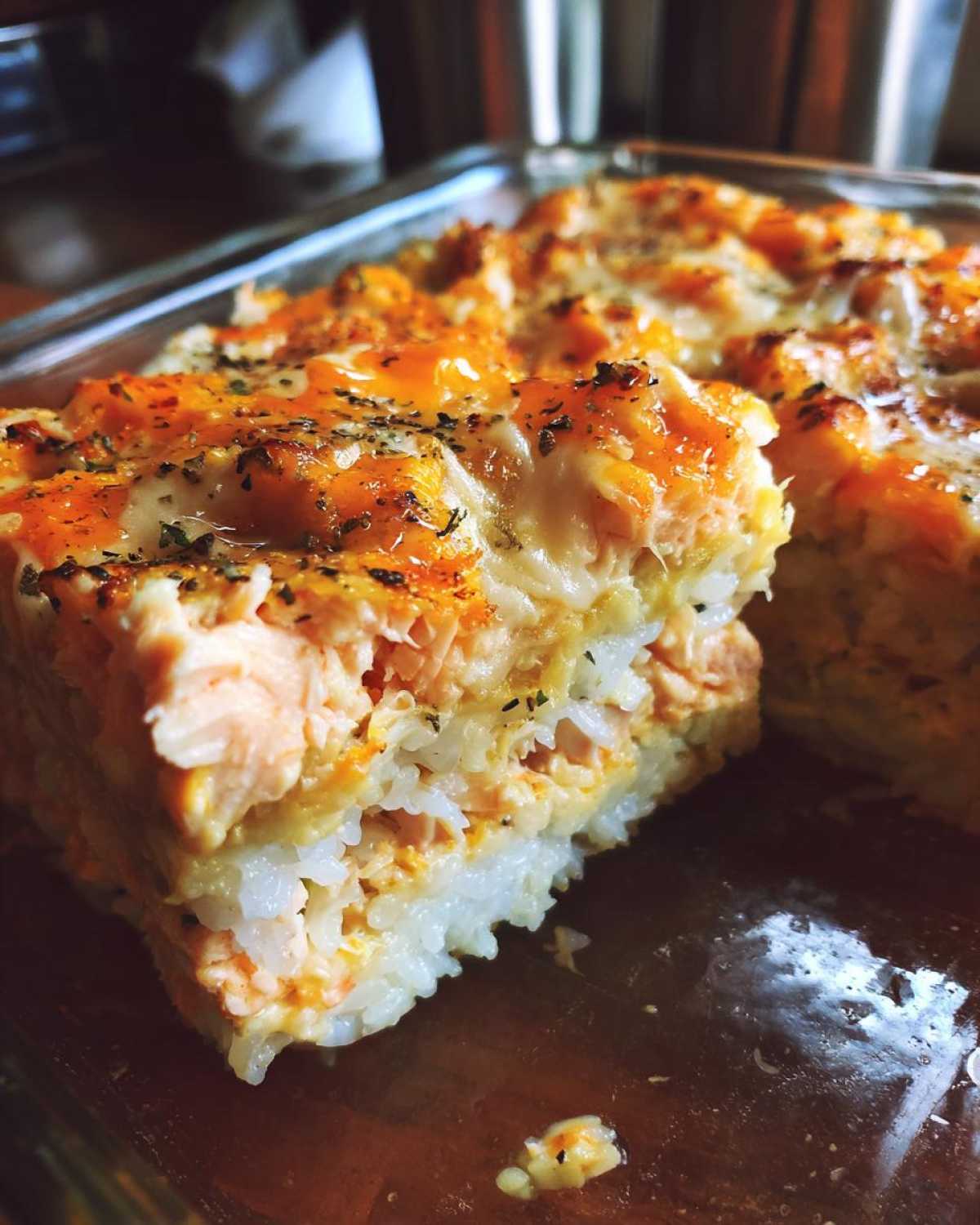 A cross-section slice of baked sushi bake showing layers of rice, salmon, and a melted, cheesy topping sprinkled with herbs.