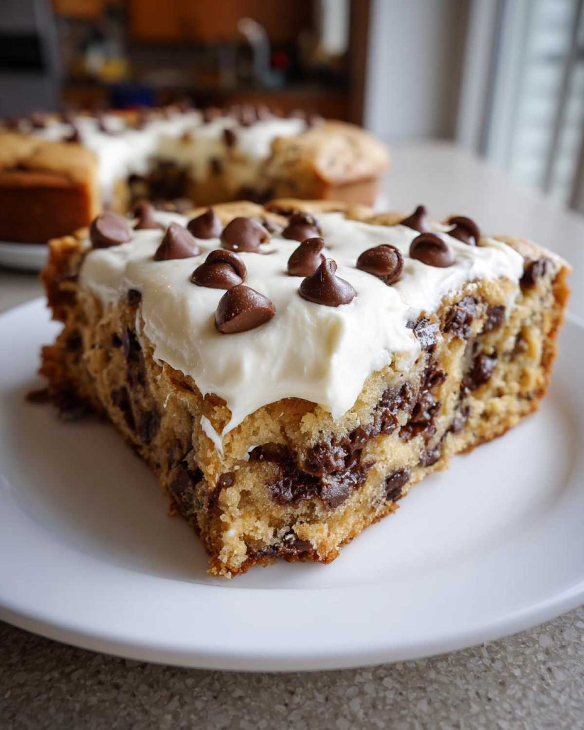 A thick slice of chewy cookie cake recipe topped with white frosting and chocolate chips.